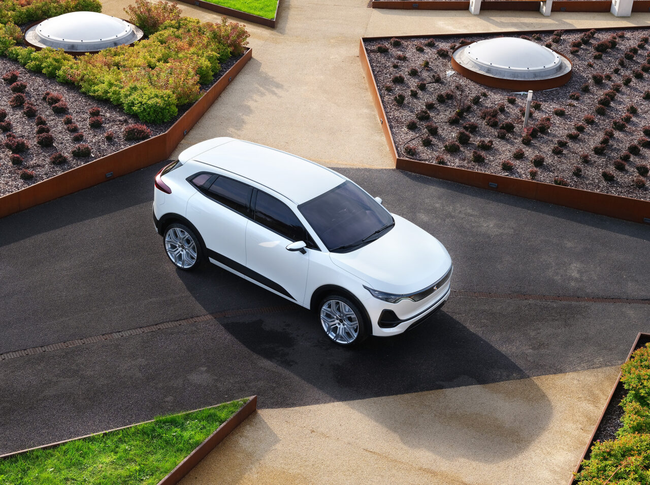 Biały samochód elektryczny Izera zaparkowany na betonowym parkingu obok zielonych dachów z roślinnością, widok z góry.