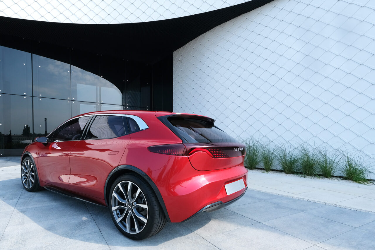 Czerwony samochód elektryczny Izera zaparkowany przed nowoczesnym oszklonym budynkiem. W tle biała elewacja i rośliny.