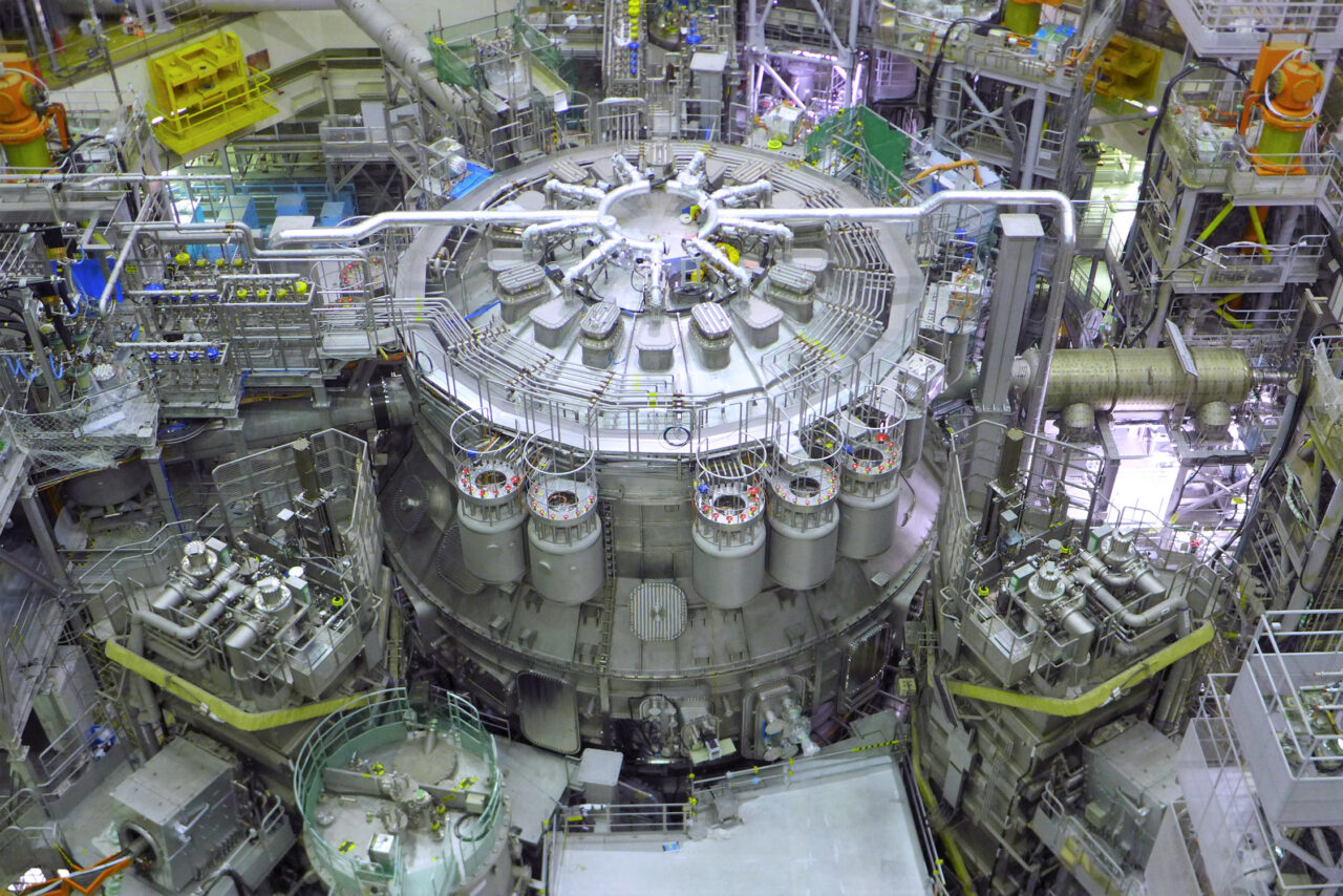 JT-60SA, największy reaktor nuklearny świata z Japonii. Instalacja przemysłowa składająca się z metalowych cylindrów i rur, rozmieszczonych symetrycznie wokół centralnej jednostki.