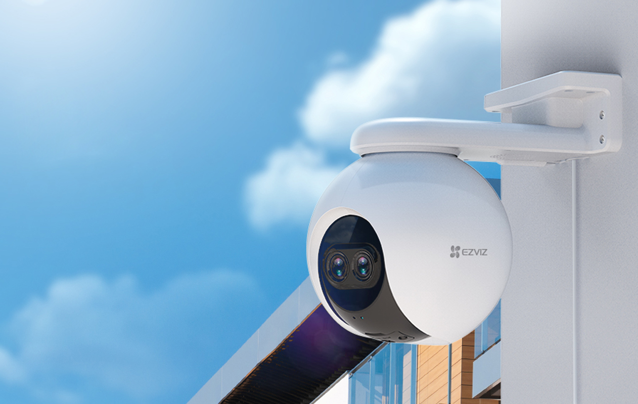 Biała kamera bezpieczeństwa Ezviz C8PF typu kopułkowego zamontowana na ścianie zewnętrznej z widokiem na niebieskie niebo z chmurami.