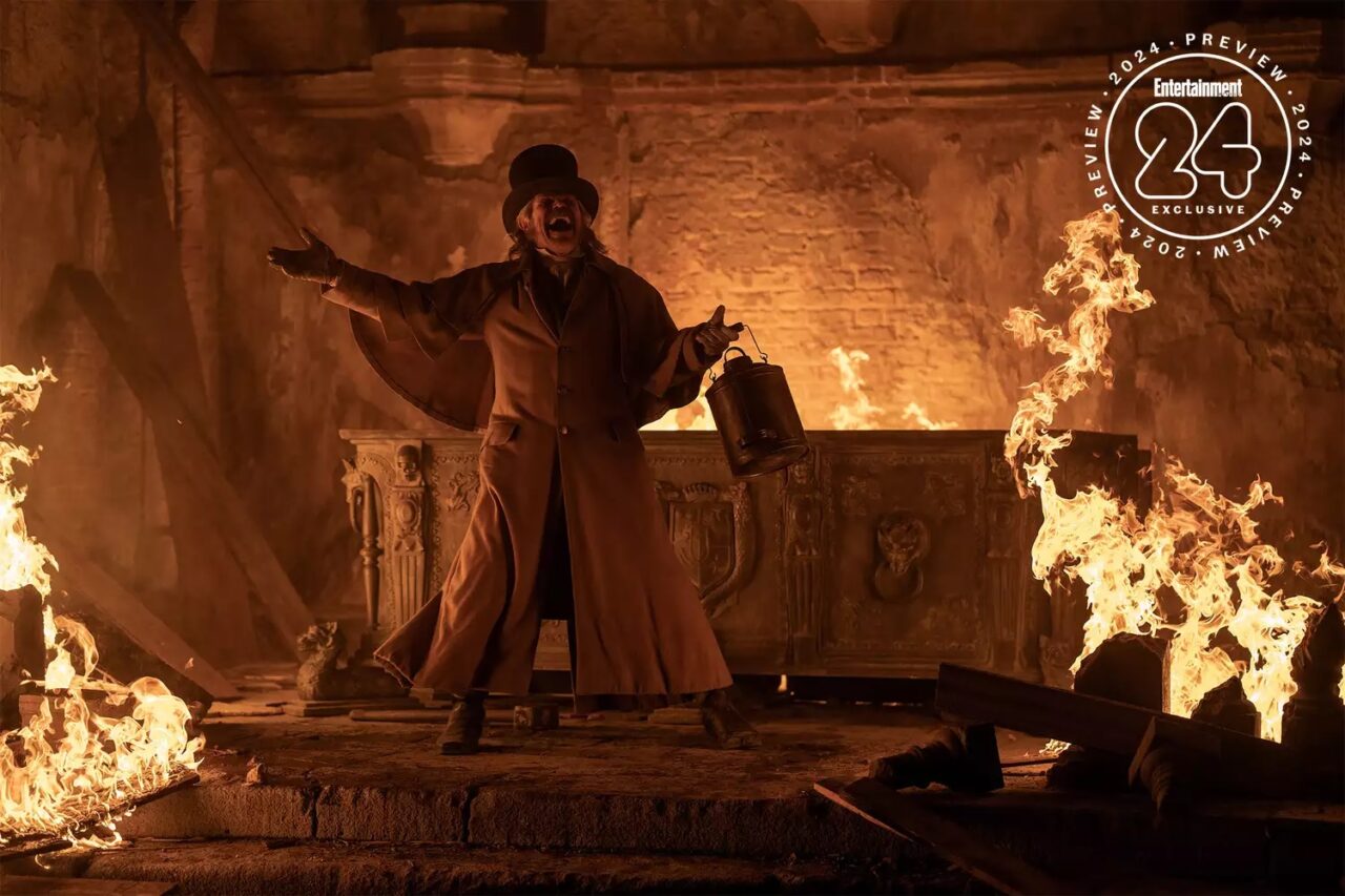 Osoba w długim płaszczu i kapeluszu stoi w centrum płonącego pomieszczenia, trzymając latarnię, z uniesionymi rękami w geście dramatycznym.