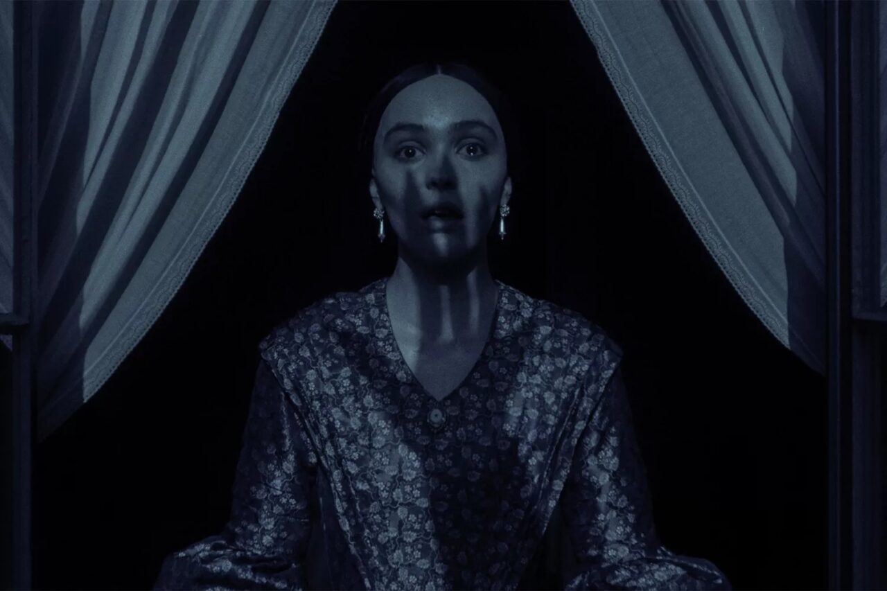 Kobieta o wyrazistej ekspresji twarzy, ubrana w tradycyjną suknię, stojąca między rozchylonymi zasłonami w zaciemnionym pomieszczeniu.