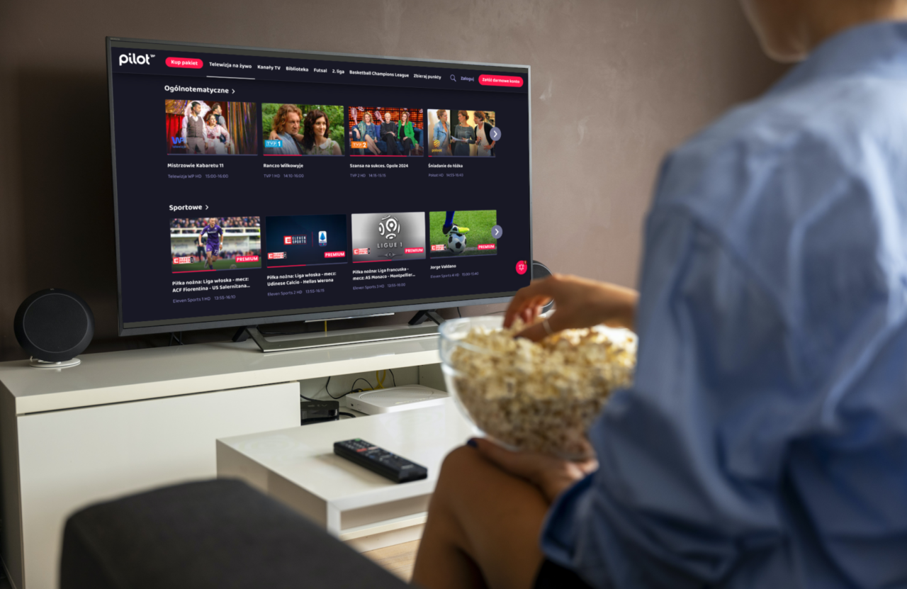 Osoba trzymająca miskę z popcornem ogląda telewizor, na którym wyświetlony jest interfejs strony pilot wp z opcjami programów telewizyjnych.