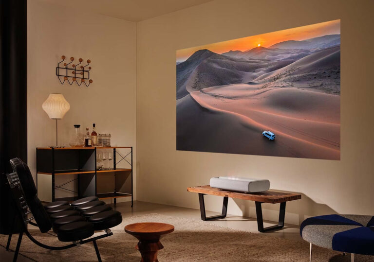 Elegancko urządzony pokój z dużym obrazem pustyni na ścianie, który rzutowany jest z projektora Samsung LSP7T The Premiere, stylową lampą, półką z alkoholem i szklanymi karafkami oraz nowoczesnymi meblami w tym leżanką i drewnianym stołkiem.