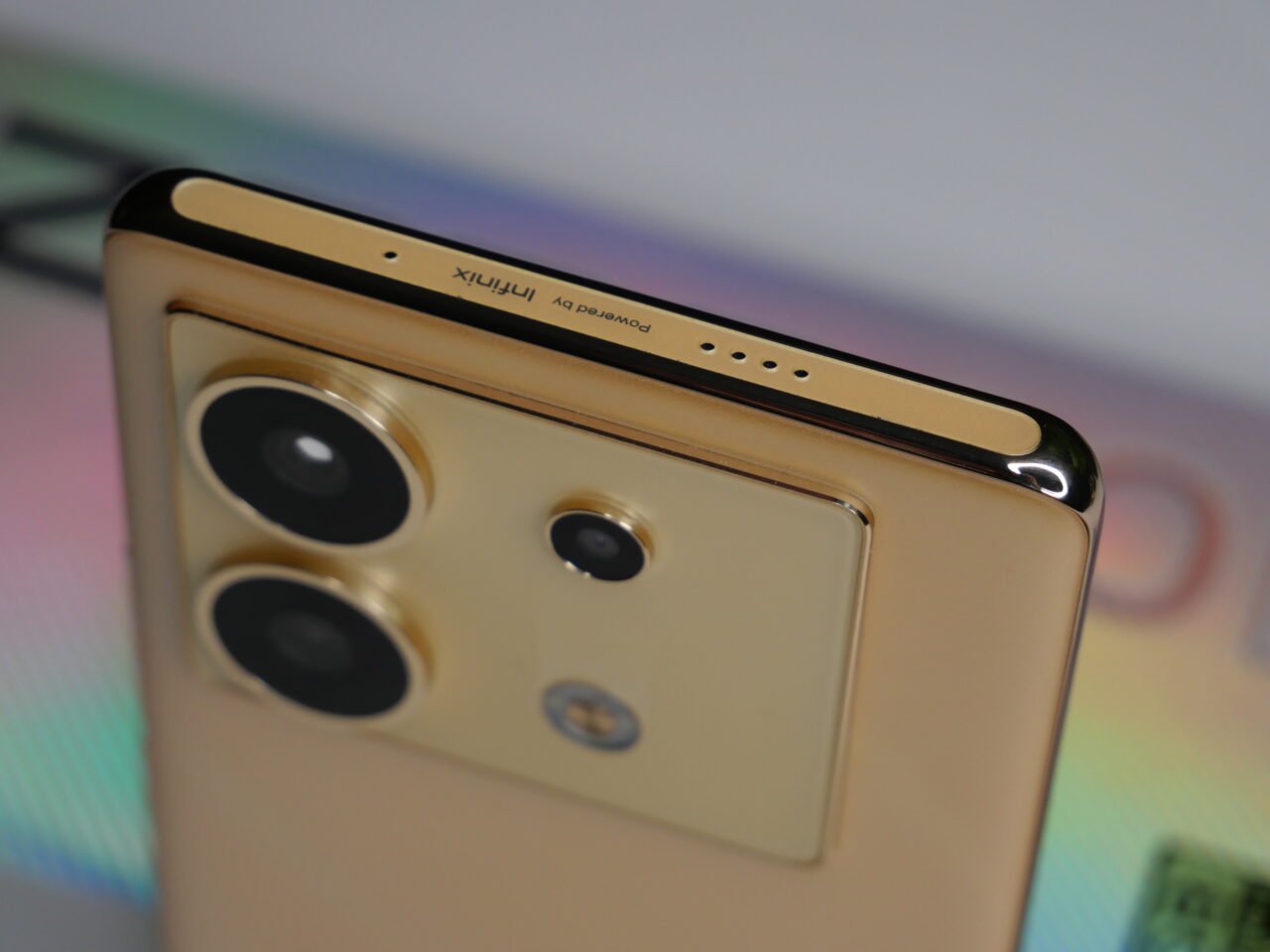 Zbliżenie na górną część złotego smartfona z potrójnym aparatem i logo producenta, przeciw tęczowemu tłu.