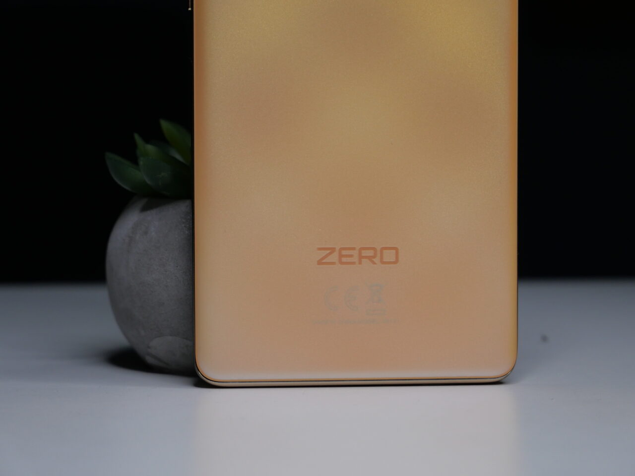Tylna część złotego smartfona z napisem "ZERO" i symbolami certyfikatów, po lewej stronie doniczka z zielonym sukulenterm, na tle czarno-białej kompozycji.