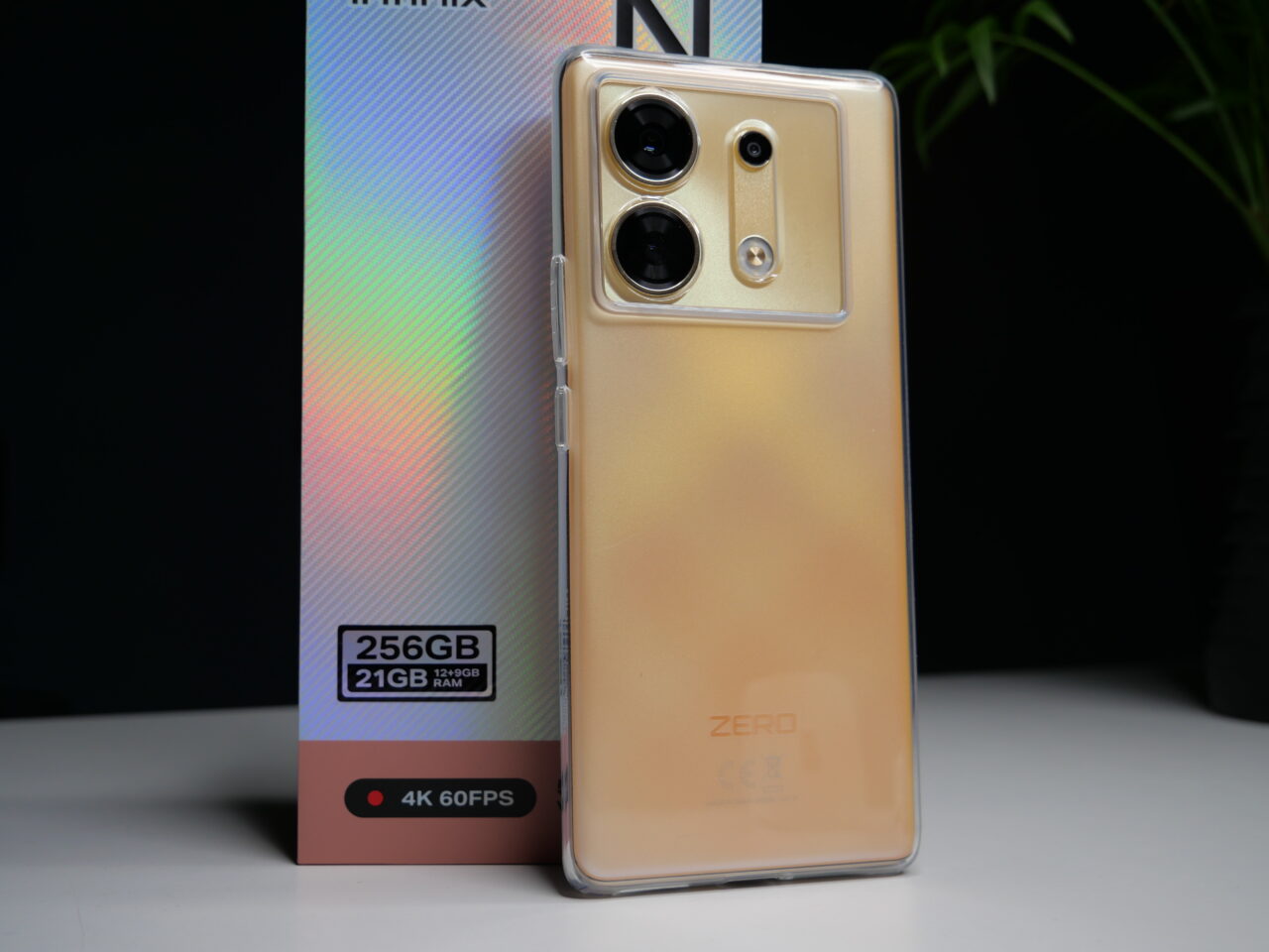 Złoty smartfon z potrójnym aparatem i błyszczącym gradientem na tylnej obudowie, oparty o pudełko z napisem "256GB 21GB RAM 4K 60FPS".