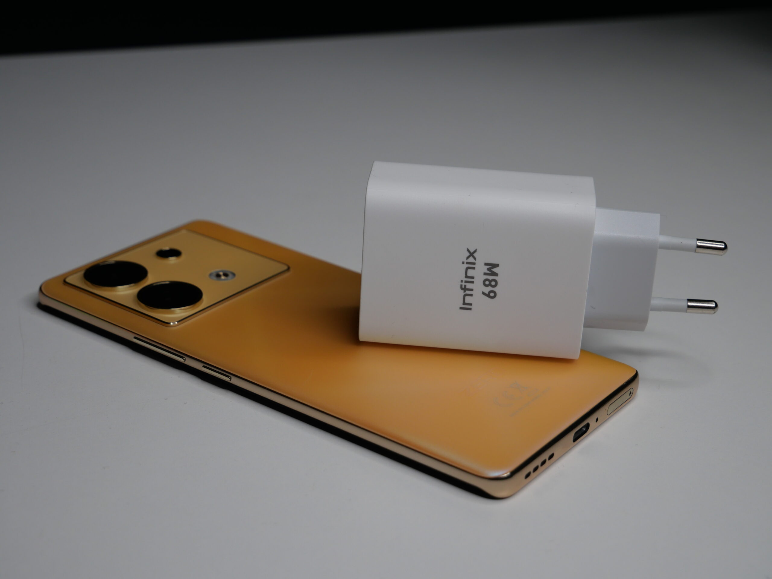 Złoty smartfon leżący na płasko z widocznymi potrójnymi aparatami z tyłu i białą ładowarką oznaczoną jako "Infinix 68W" umieszczoną na jego ekranie.