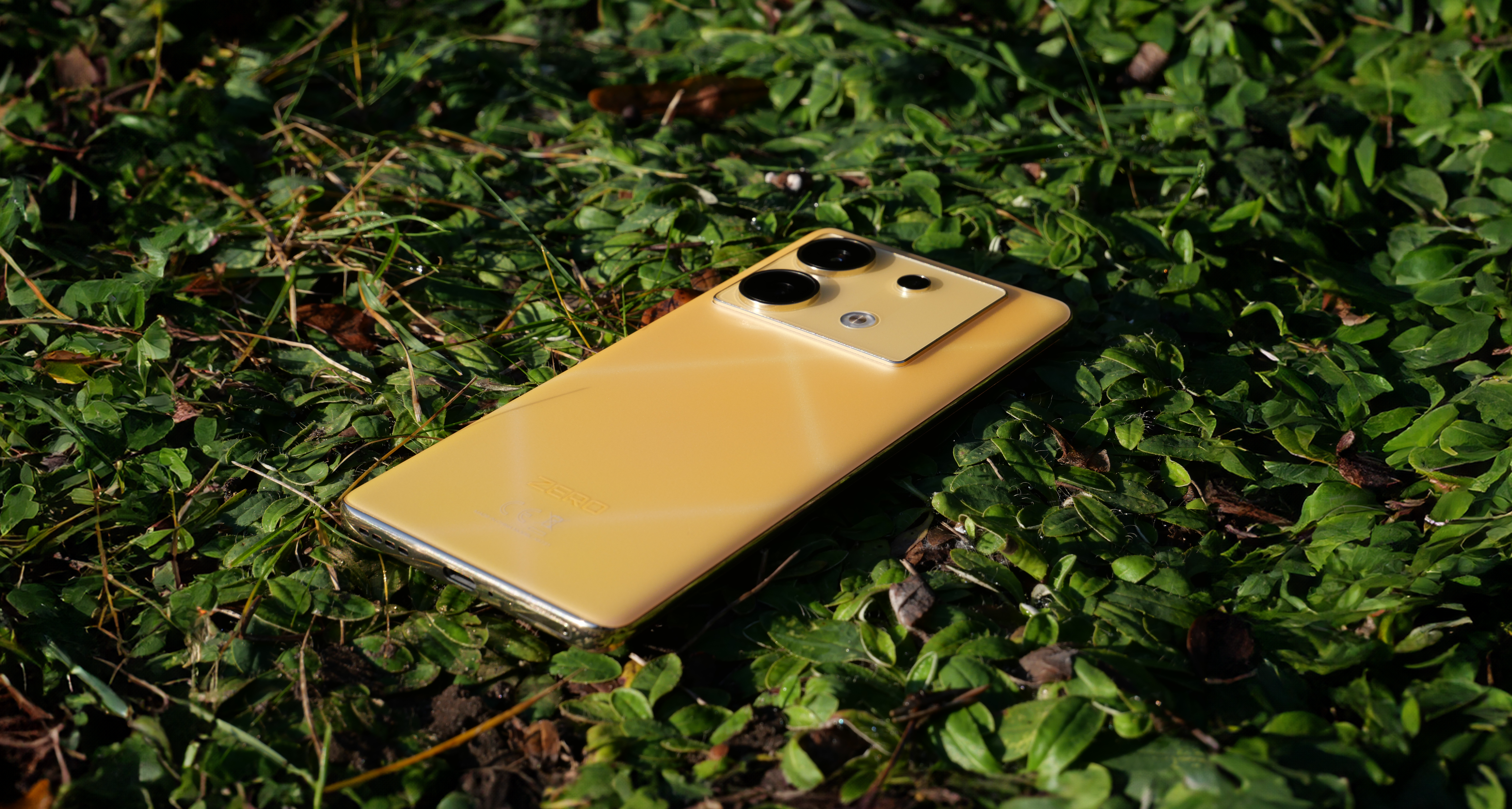 Recenzowany smartfon Infinix Zero 30 5G w złotym kolorze leżączy na zielonych, małych roślinach