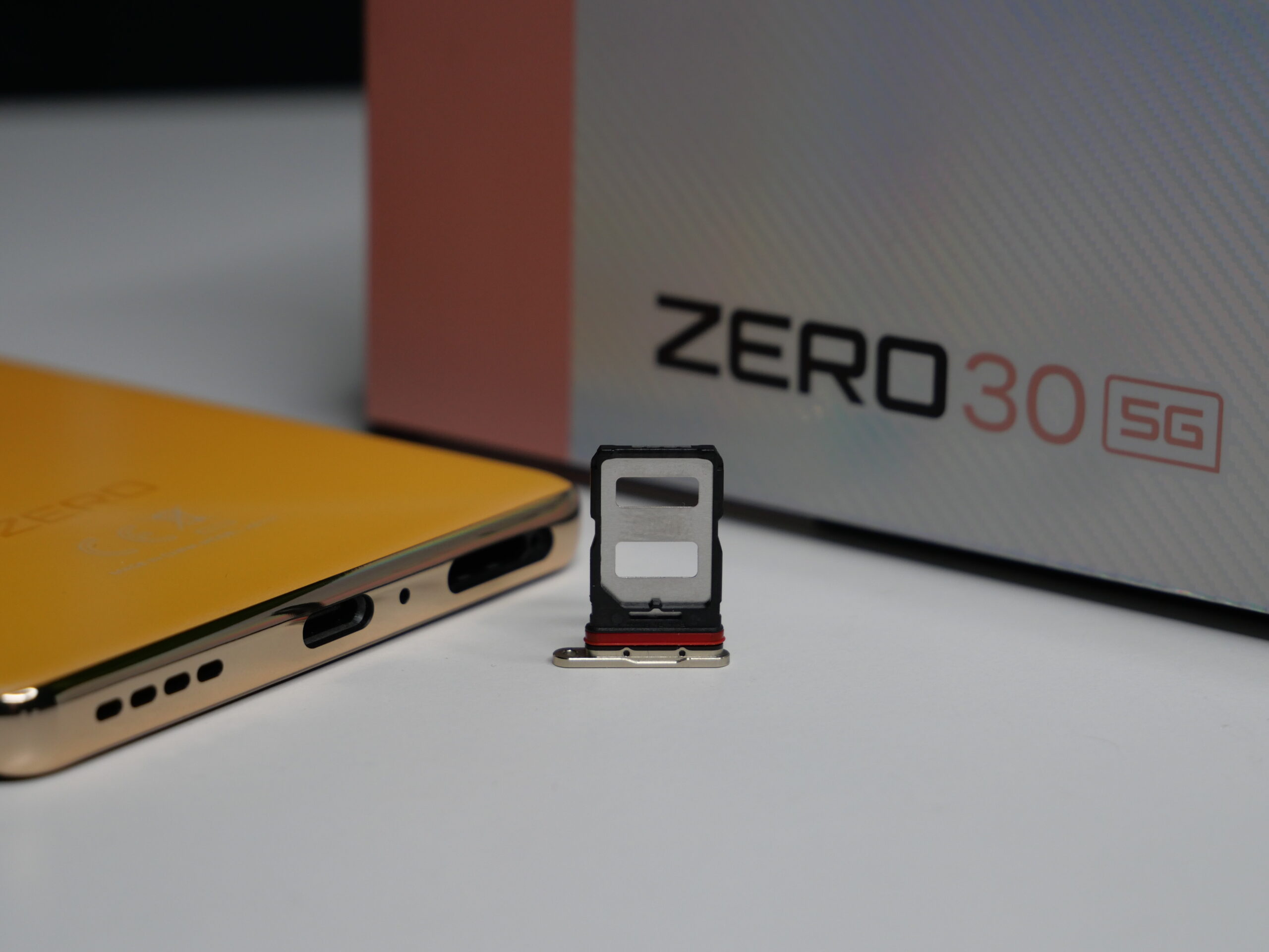 Złoty smartfon z napisem "ZERO" położony na białej powierzchni obok otwartej tacki karty SIM, w tle pudełko z napisem "ZERO 30 5G".