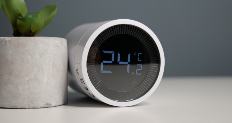 Cyfrowa głowica termostatyczna tesla o okrągłym kształcie pokazujący temperaturę 24,5°C. Obok betonowej doniczki z rośliną.
