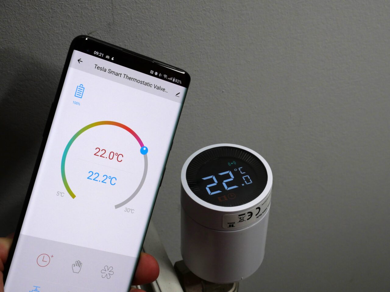 Smartfon wyświetlający aplikację do zarządzania temperaturą z czytelnym termometrem pokazującym 22,0°C, obok inteligentnego termostatu naściennego również pokazującego 22°C.