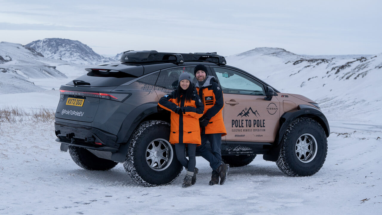Mężczyzna i kobieta w pomarańczowych kurtkach stoją uśmiechnięci. Z tyłu znajduje się samochód elektryczny z oznaczeniami ekspedycji "Pole to Pole" na tle zimowego krajobrazu z pokrytymi śniegiem wzgórzami.