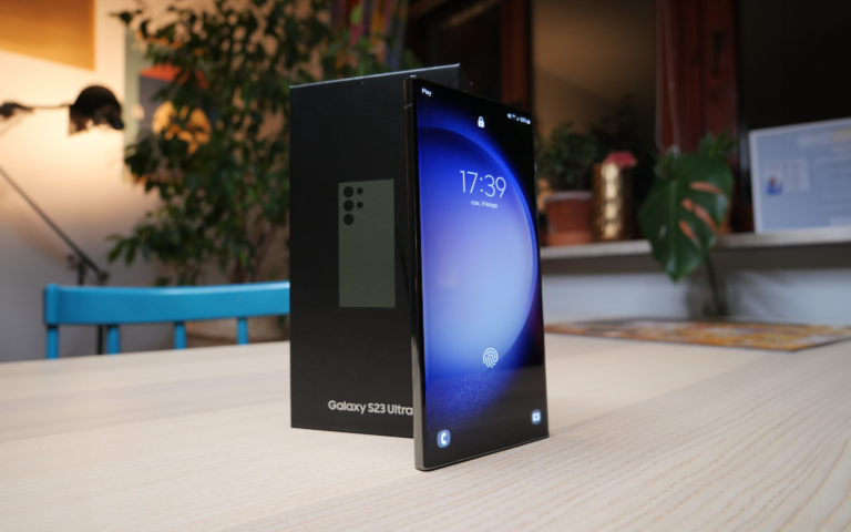 Smartfon Galaxy S23 Ultra stojący pionowo na drewnianym biurku obok swojego opakowania, z wyświetlaczem pokazującym ekran blokady i godzinę. W tle rozmyte wnętrze domu.