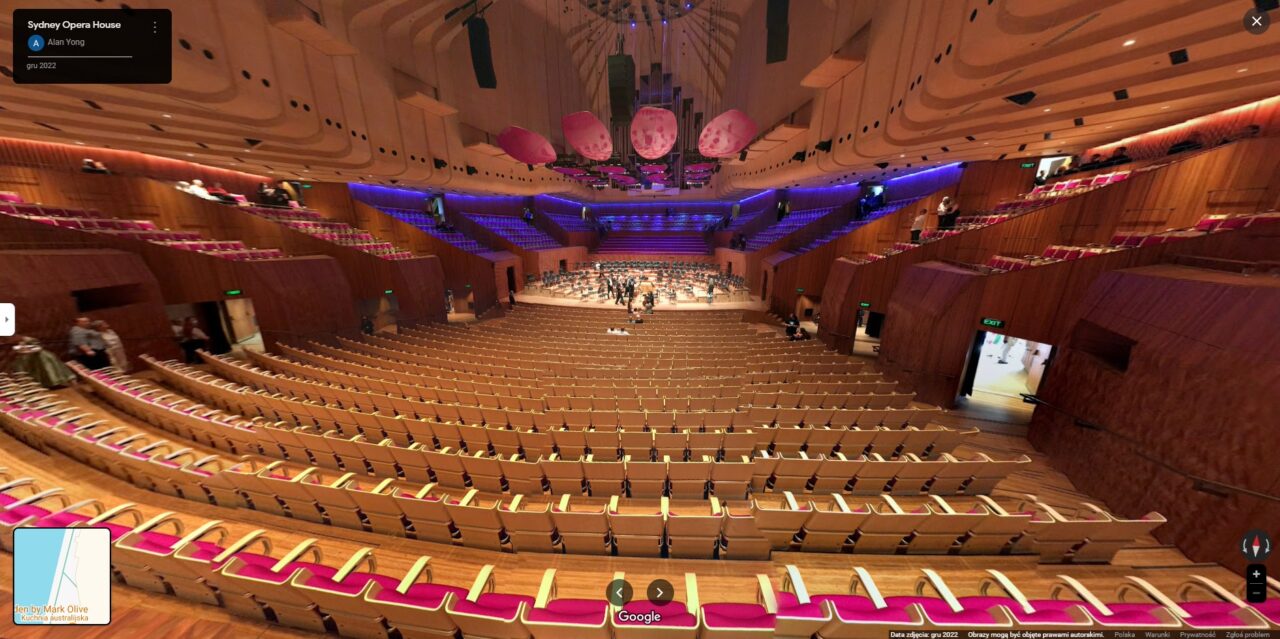 Wnętrze Opery w Sydney z widocznymi rzędami foteli w kolorze beżowym i fuksji oraz sceną z ludźmi w tle.