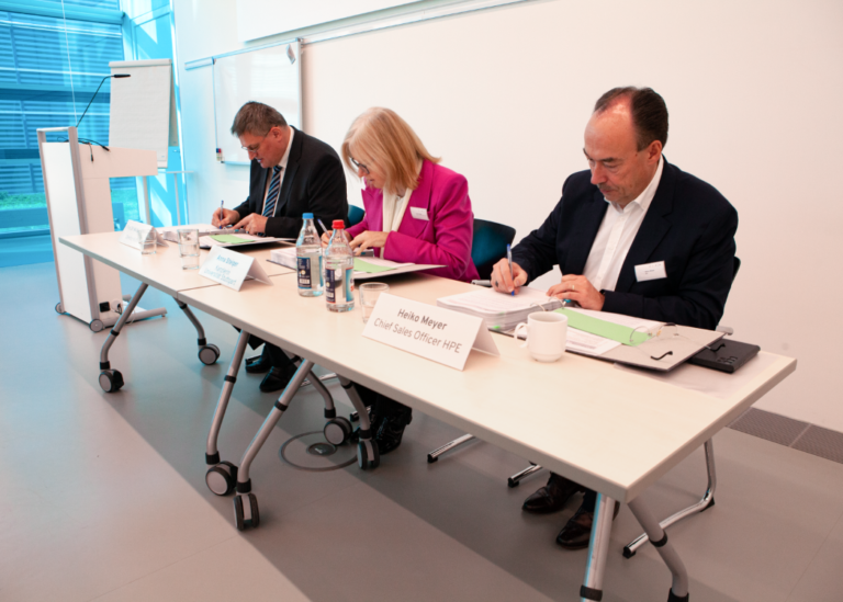 Trzy osoby siedzące przy długim biurku i piszące na dokumentach w jasnym pomieszczeniu konferencyjnym; na pierwszym planie widoczne tabliczki z nazwiskami i stanowiskami.