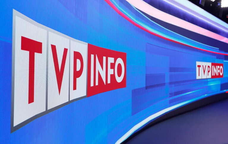 Wyświetlacz z logo TVP Info w studio telewizyjnym, z niebiesko-czerwonymi kolorami w tle.