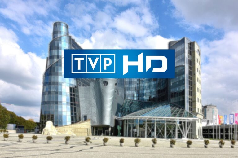 Siedziba Telewizji Polskiej z logiem TVP HD na pierwszym planie, nowoczesne budynki z przeszklonymi fasadami, przed budynkiem plac z rozmieszczonymi donicami z krzewami.