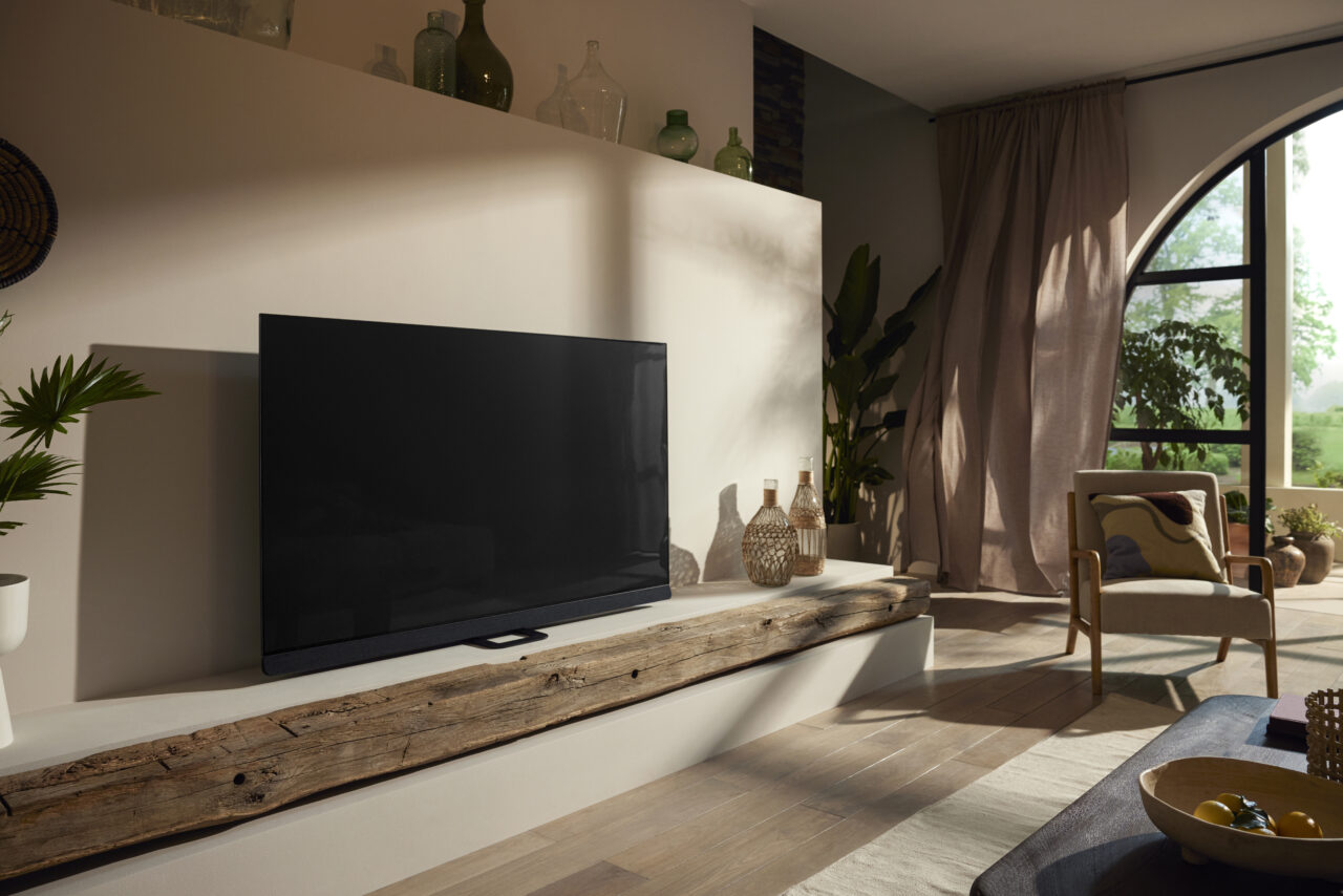 Abonament RTV. Stylowy salon z dużym telewizorem Philips Ambilight OLED+908 na drewnianej półce, wygodnym fotelem przy oknie i dekoracyjnymi butelkami na półce nad telewizorem.