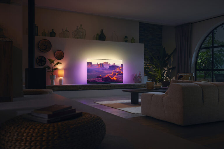Nowoczesny salon z włączonym telewizorem Philips Ambilight OLED+908, wyświetlającym obraz skalistej pustyni, otoczonego różowym oświetleniem LED. W tle eleganckie, ciemne meble i rośliny domowe.