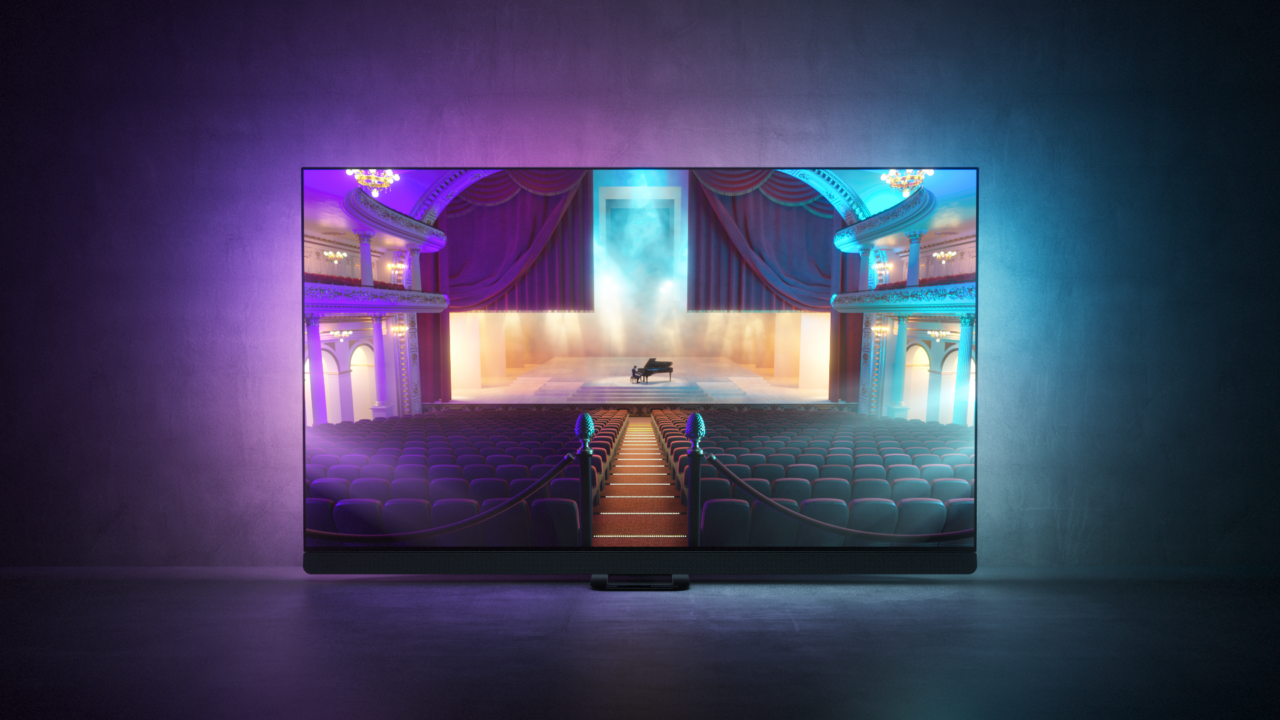 Telewizor w ciemnym pomieszczeniu wyświetlający obraz wnętrza teatru z czerwonymi zasłonami, balkonami i fortepianem na scenie.