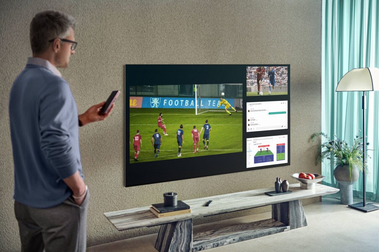 Mężczyzna w okularach stoi w salonie z pilotem w ręku i patrzy na wiszący na ścianie duży telewizor Samsunga, na którym wyświetlany jest mecz piłki nożnej oraz interfejs użytkownika z komentarzami i statystykami.