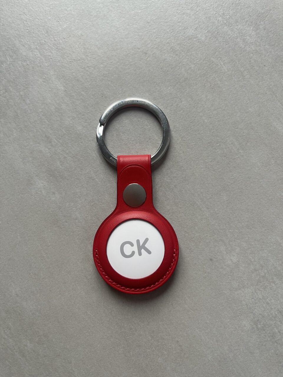 Czerwony AirTag z metalowym kółkiem i okrągłym przywieszkiem z białą wstawką z napisem "CK".