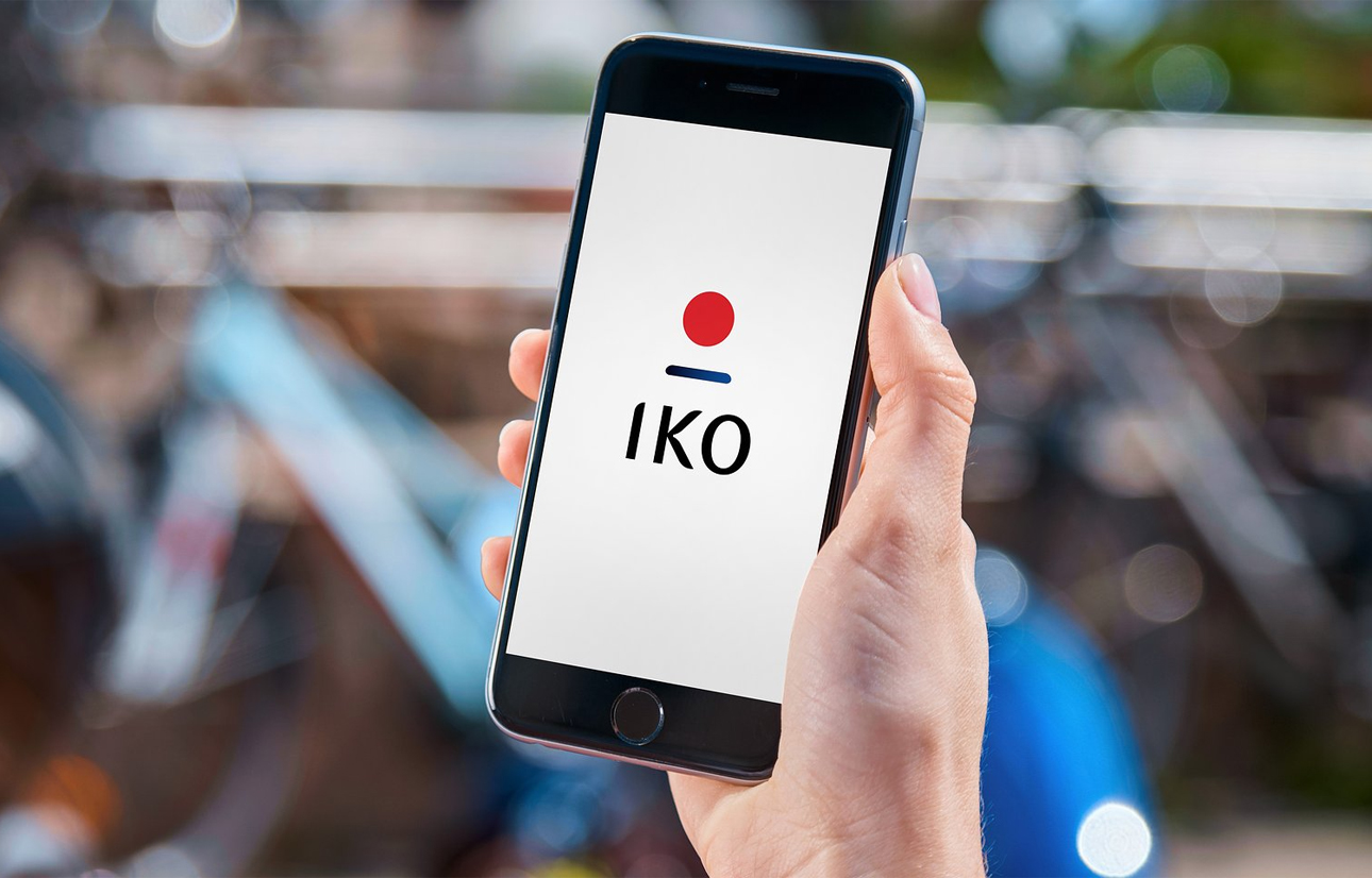 Ręka trzymająca smartfon z logo IKO na ekranie na rozmytym tle miejskiego krajobrazu.