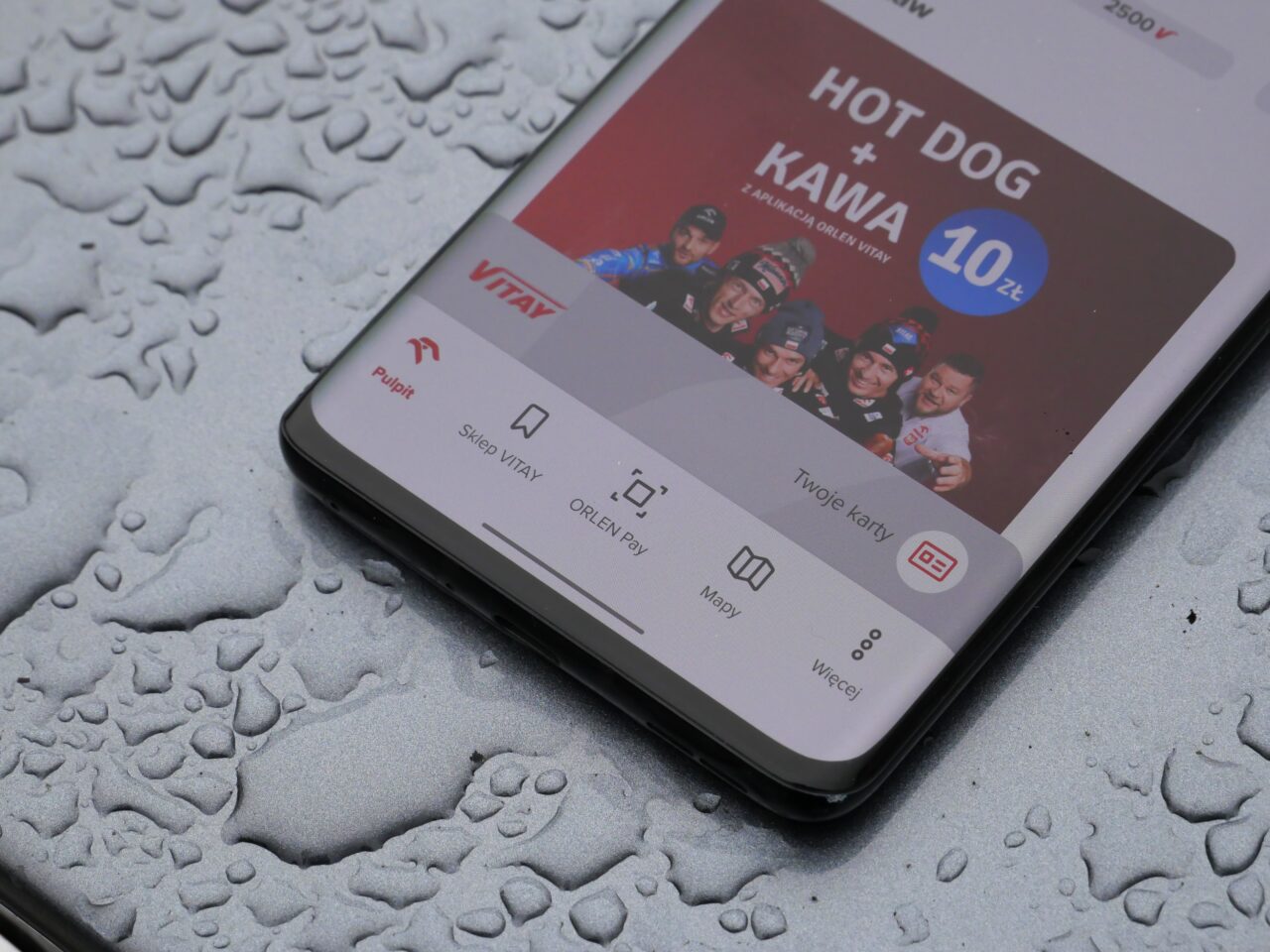 Smartfon leżący na mokrej powierzchni wyświetla reklamę z grupą osób i tekstem "HOT DOG + KAWA 10 zł" na ekranie.