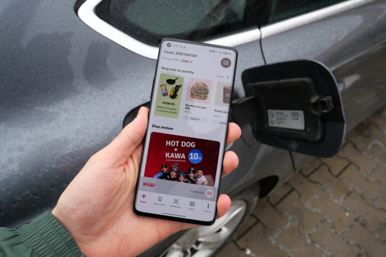 Dłoń trzymająca smartfon z otwartą aplikacją Orlen VITAY przed szarym samochodem z otwartą klapką wlewu paliwa.