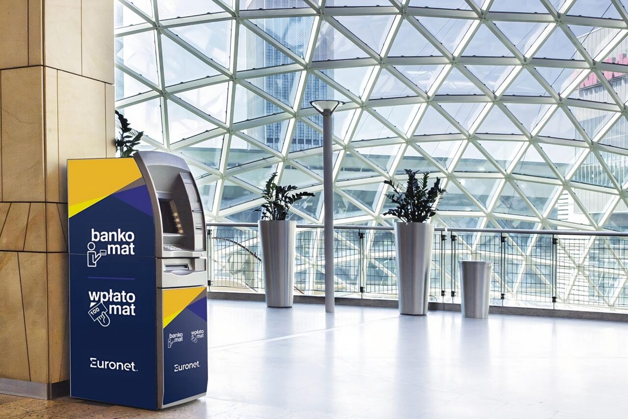 Bankomat Euronet w nowoczesnym wnętrzu ze szklanym dachem i donicami z roślinami.
