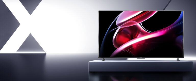 Nowoczesny telewizor stojący na minimalisticznym stoliku w eleganckim wnętrzu z dużą czarno-białą literą "X" w tle. Na ekranie wyświetlany jest wizualny obraz wyglądający jak kolorowe płynne figury w odcieniach różu i błękitu.