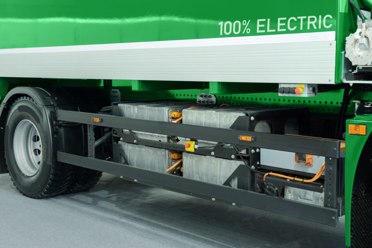 Zielony elektryczny pojazd ciężarowy z widocznymi bateriami i komponentami elektrycznymi na boku, z napisem "100% ELECTRIC".