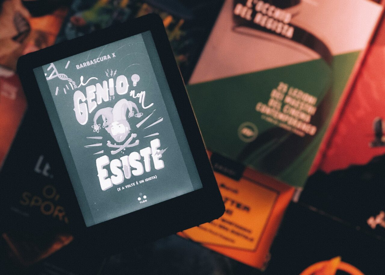 Czytnik e-booków wyświetlający okładkę książki z napisem "Genio non esiste", otoczony fizycznymi książkami w tle.