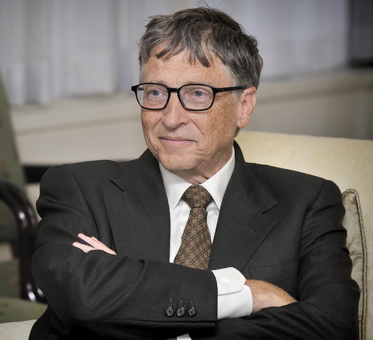 Bill Gates w średnim wieku z okularami, w eleganckim garniturze, siedzi z założonymi rękami i lekko się uśmiecha.
