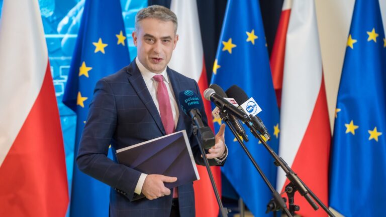 minister cyfryzacji Krzysztof Gawkowski stojący przed mikrofonami na tle flag Polski i UE, bez AI