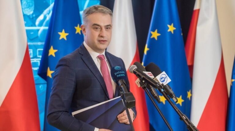 minister cyfryzacji Krzysztof Gawkowski stojący przed mikrofonami na tle flag Polski i UE