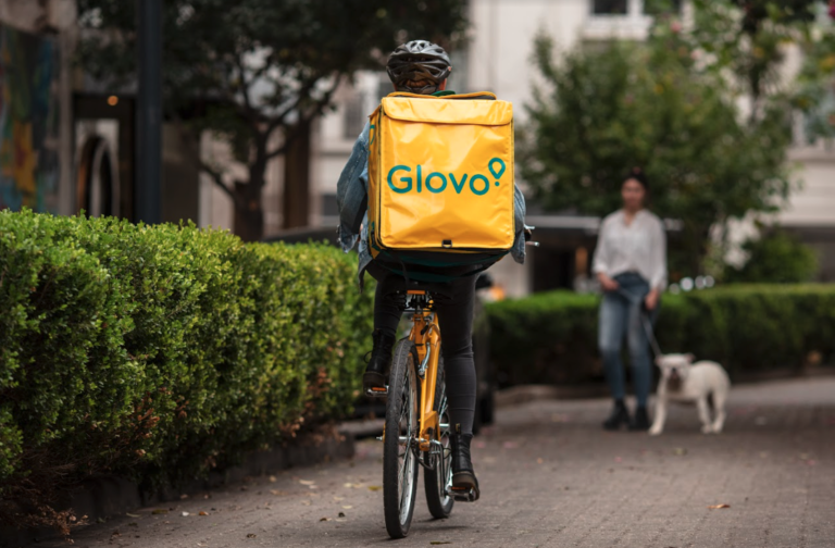 Dostawca Glovo na rowerze z żółtym plecakiem firmy Glovo jedzie ulicą; w tle widać spacerującą osobę z psem.