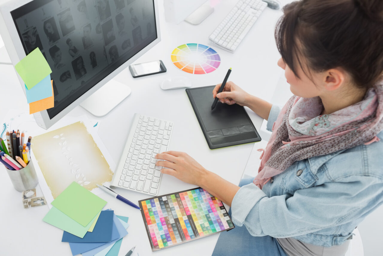 Graficzka pracuje przy biurku z użyciem tabletu graficznego, monitora, klawiatury i kółka barw.