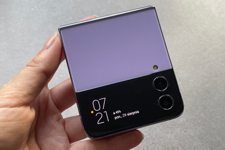 Zdjęcie ręki trzymającej złożony smartfon Samsung Galaxy Z Flip 4 z systemem One UI 6.0 z zewnętrznym wyświetlaczem pokazującym godzinę, datę i poziom naładowania baterii.