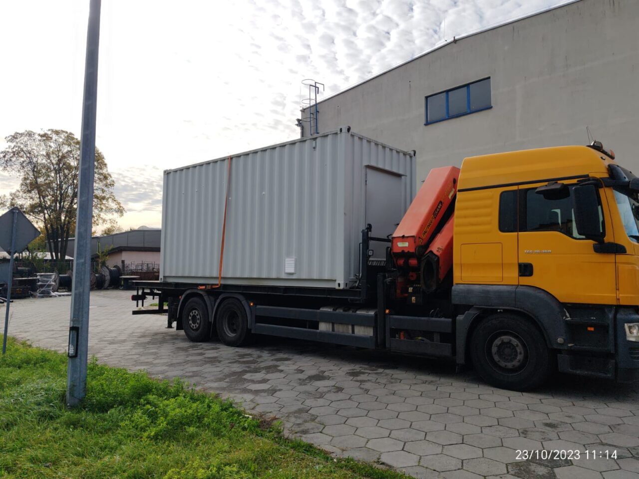 Ciężarówka z żółtą kabiną i kontenerem na naczepie stoi na brukowanym parkingu przy budynku przemysłowym.