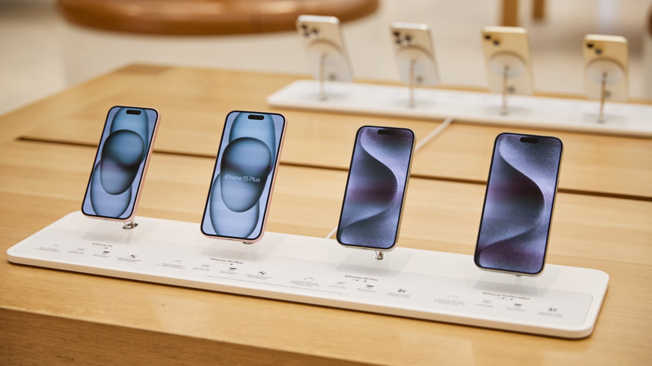 Cztery smartfony ustawione na wystawie w sklepie, z etykietami modeli i specyfikacji na dole.
