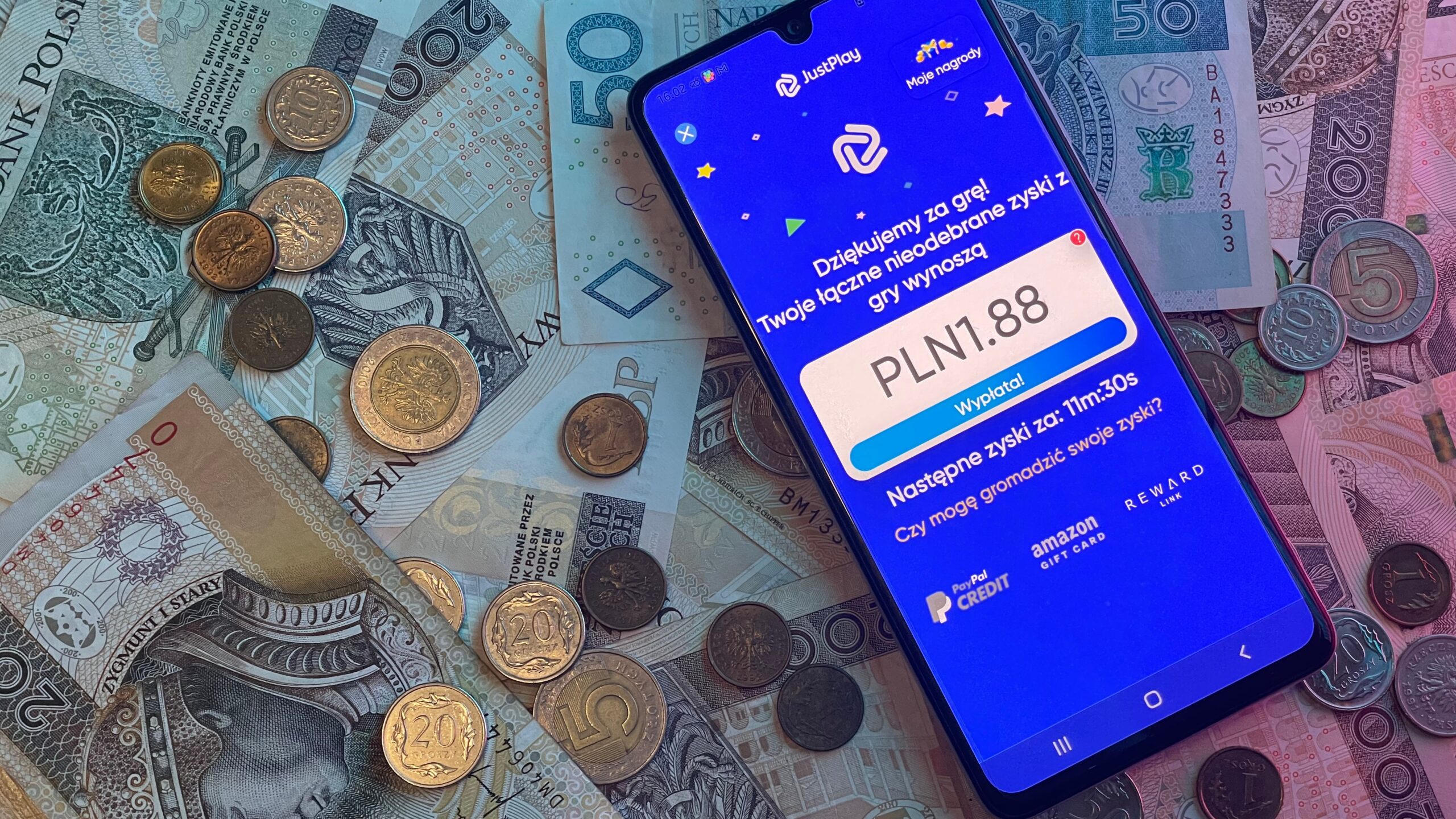 Smartfon leżący na polskich banknotach i monetach z otwartą aplikacją mobilną, która wyświetla komunikat o sumie wygranej 1,88 PLN.