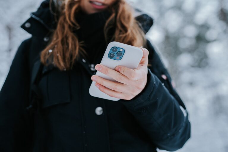 Kobieta w czarnej kurtce trzymająca smartfon iPhone w etui z widocznym aparatem, w tle zimowy krajobraz.