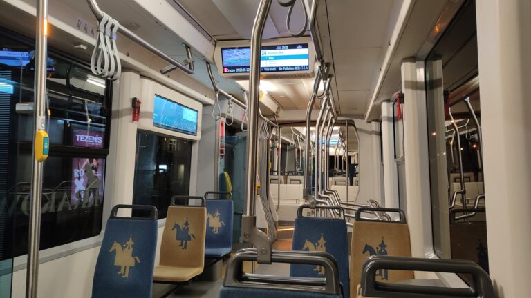 Kraków. Wnętrze pustego tramwaju wieczorem z niebieskimi i beżowymi siedzeniami oraz metalowymi uchwytami dla pasażerów. Na wyświetlaczach informacyjnych widoczne aktualne informacje dotyczące jazdy.