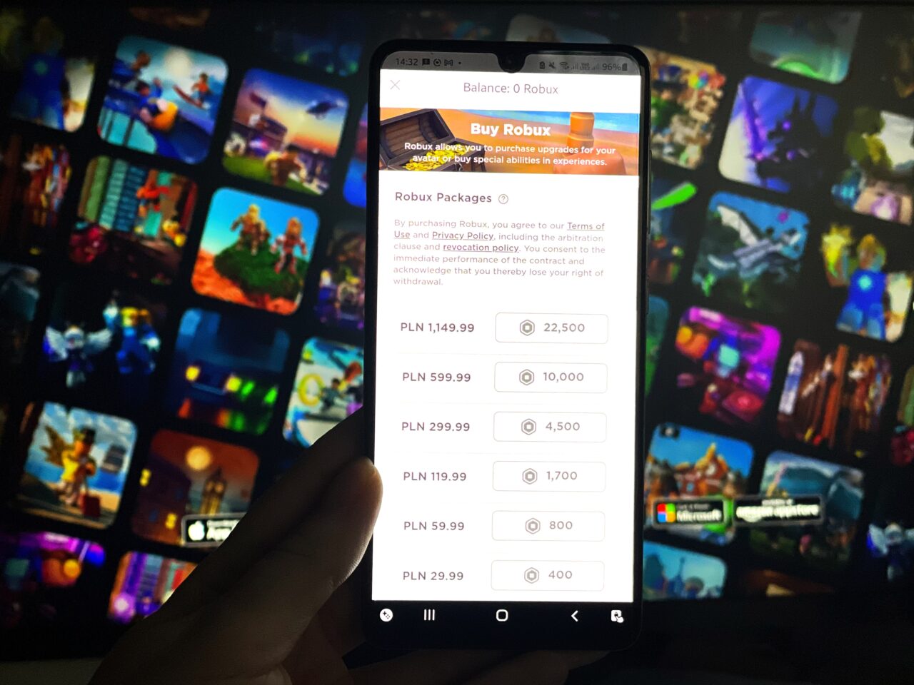 Dłoń trzymająca smartfon z otwartą aplikacją oferującą zakup waluty wirtualnej Robux w grze Roblox, z rozmytym tłem pokazującym barwne ekranowe sceny z gier wideo.