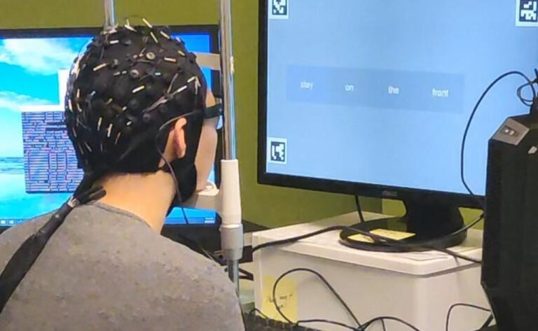 Osoba z elektrodami EEG na głowie patrzy na monitor komputera, na którym wyświetlony jest eksperyment z użyciem bodźców wizualnych. Testuje ona sterowanie myślami
