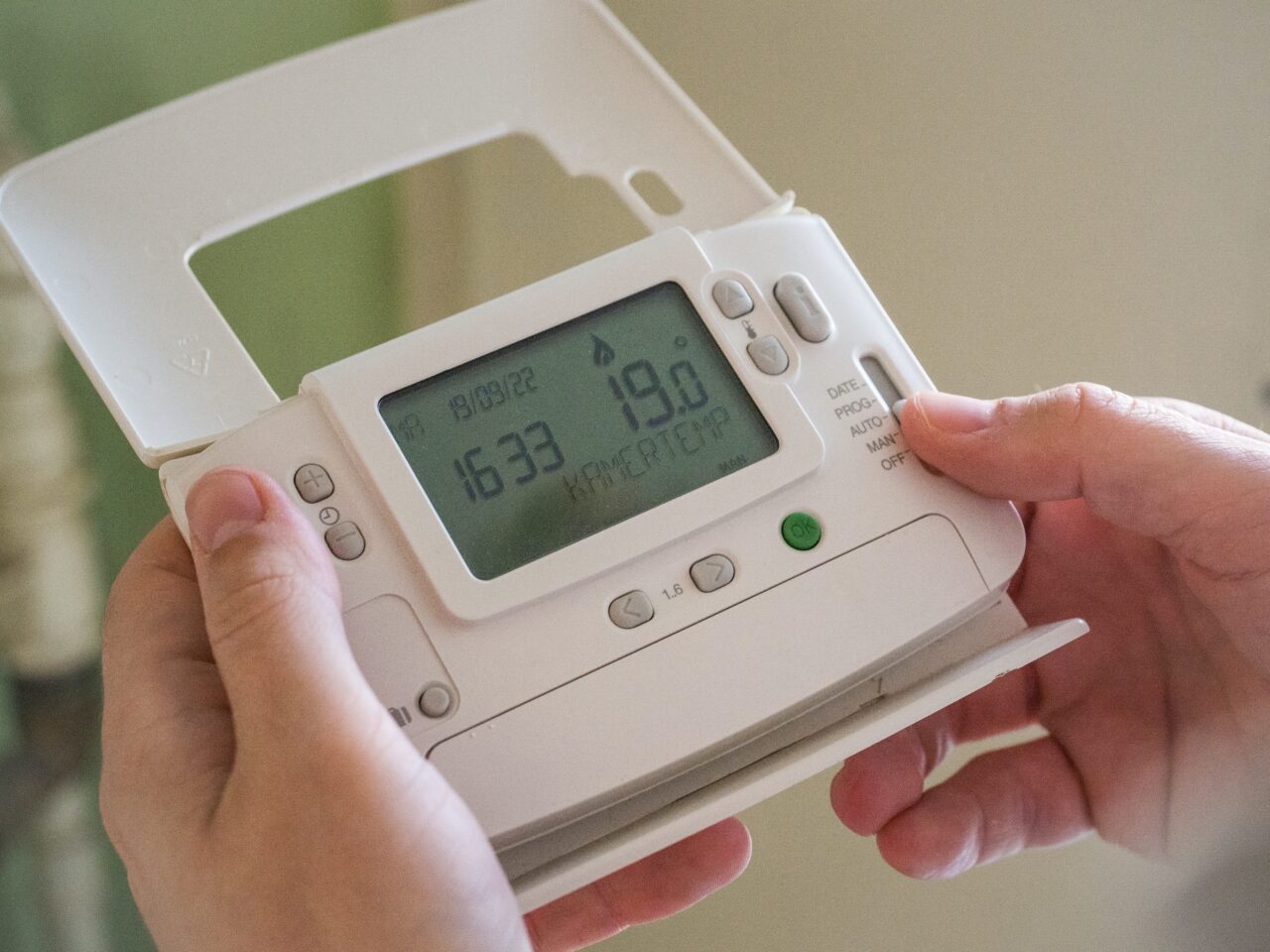 Osoba trzymająca w dłoniach biały, cyfrowy termostat pokojowy wyświetlający godzinę i temperaturę.
