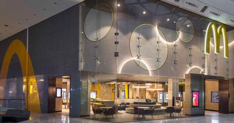 Wnętrze restauracji szybkiej obsługi z charakterystycznym złotym logo McDonald's, nowoczesnym wystrojem oraz stolikami i krzesłami dla klientów.