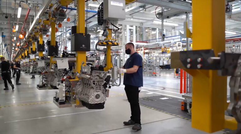 Pracownik w masce stoi obok zawieszonego silnika w nowoczesnej hali produkcyjnej w fabryce Mercedes z robotami przemysłowymi i montażowymi liniami w tle.